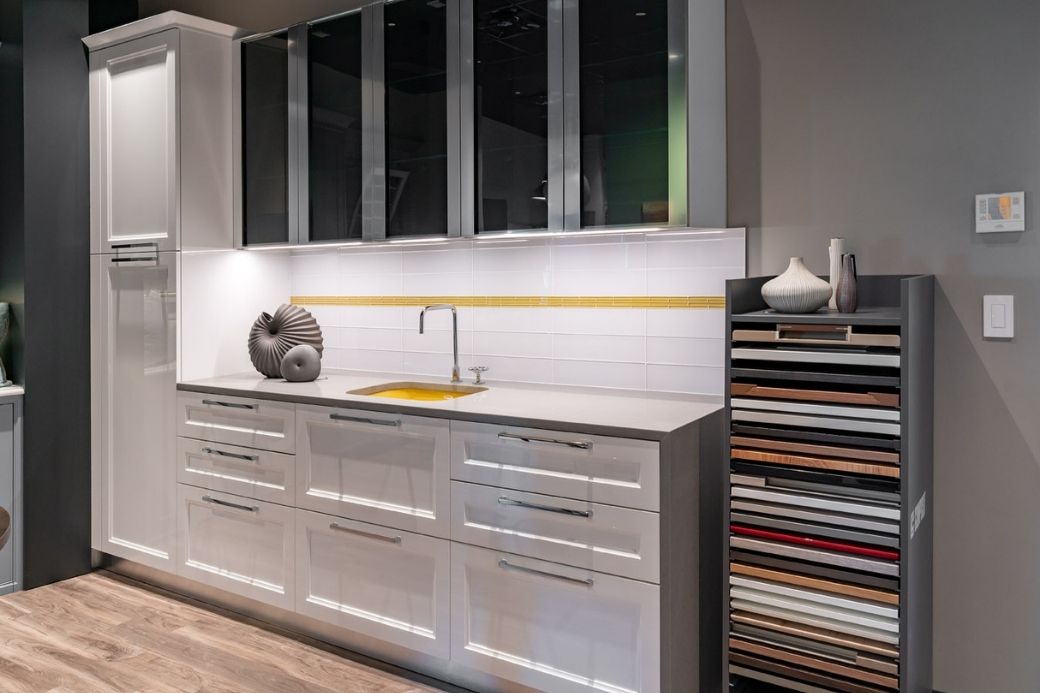 View of a sample counter space in Interiology showroom with cabinets featuring sleek grays contrasted with white tile and yellow accents. 